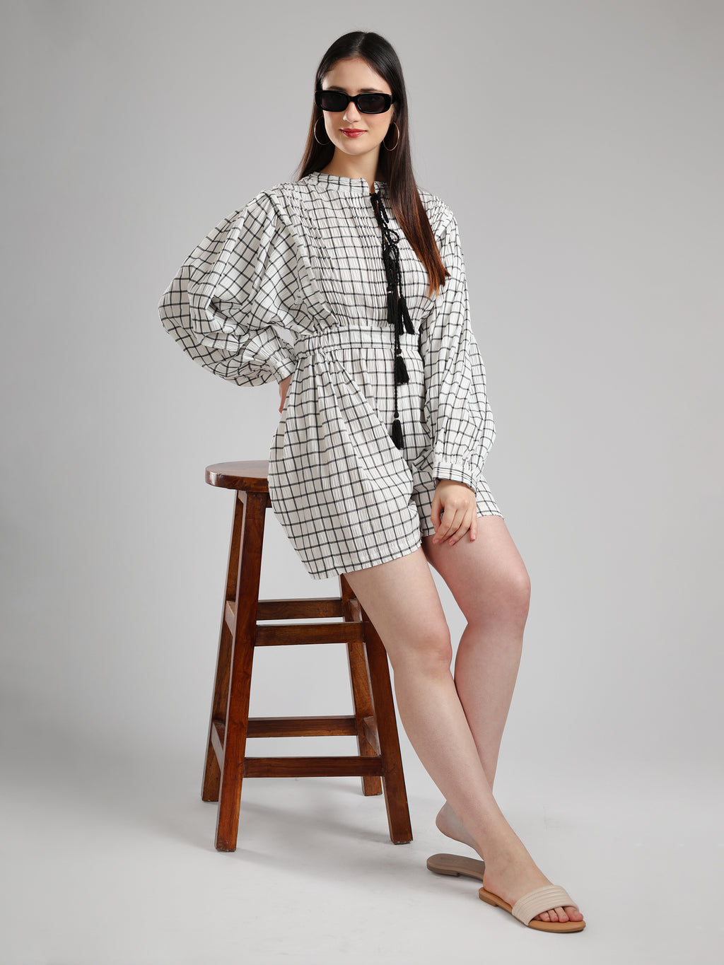 Woman wearing a black and white checkered outfit with sunglasses, standing next to a wooden stool on a gray background.