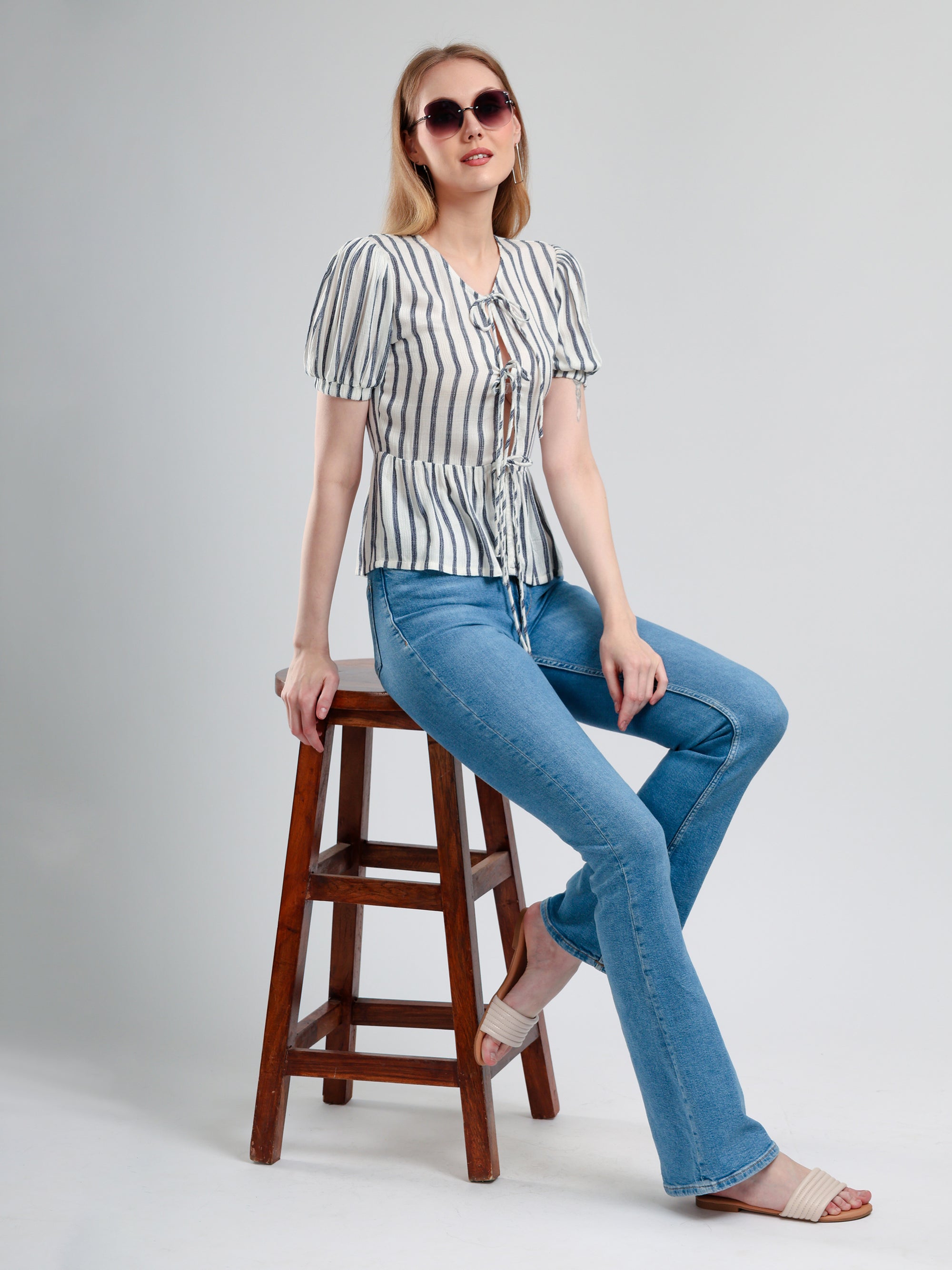 Woman sitting on a wooden stool wearing a striped shirt and blue jeans against a plain background