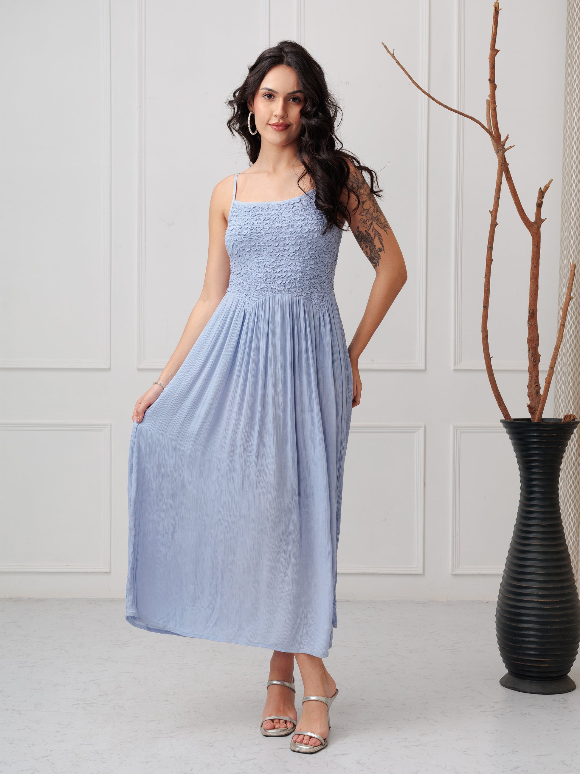 Woman wearing a light blue dress standing in a minimalistic room with a vase in the background.
